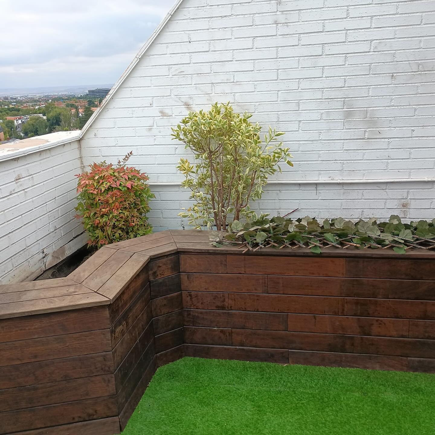 Terraza con banco de madera y jardineras, con césped artificial y pared blanca en Madrid.